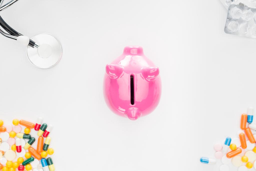 flat lay with pink piggy bank surrounded by colorful various pills, stethoscope and empty pills packages on white surface  - Photo, Image