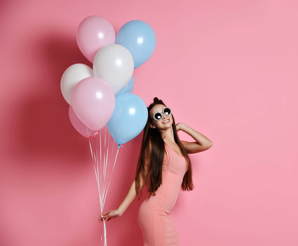 Nahaufnahme eines niedlichen blonden Mädchens, das in einem Studio steht, breit lächelt und mit blauen und rosa Luftballons spielt. - Foto, Bild