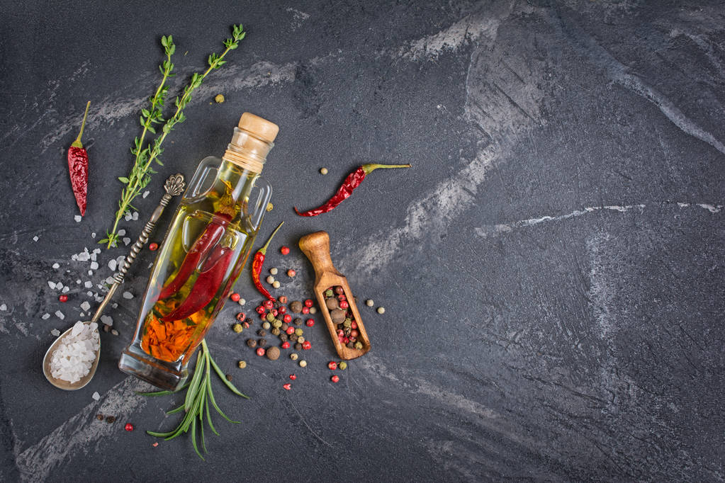 Aromatické nebo ochucený olivový olej ve skleněné láhvi s kořením a bylinkami jako chilli papričky, rozmarýn a tymián na černé mramorové pozadí s kopií prostor - Fotografie, Obrázek