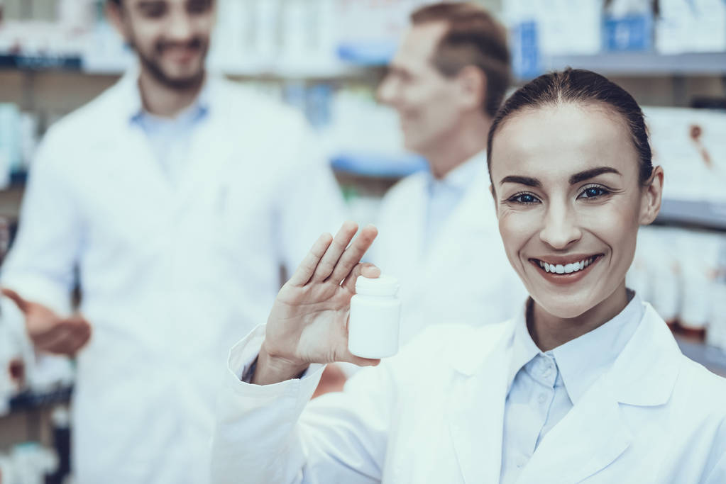 Pharmacists is Working. Pharmacists is a Caucasian Men and Woman. Woman Showing a Bottle with Pills. Men Talking on Background. People Wearing a Special Medical Uniform. People Located in Pharmacy. - Photo, Image