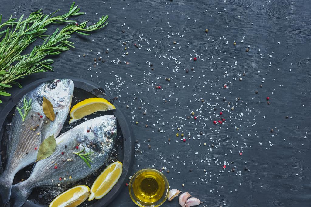 view from above of raw fish with lemon, bay leaves and rosemary in tray on black table covered by salt and pepper - Photo, Image