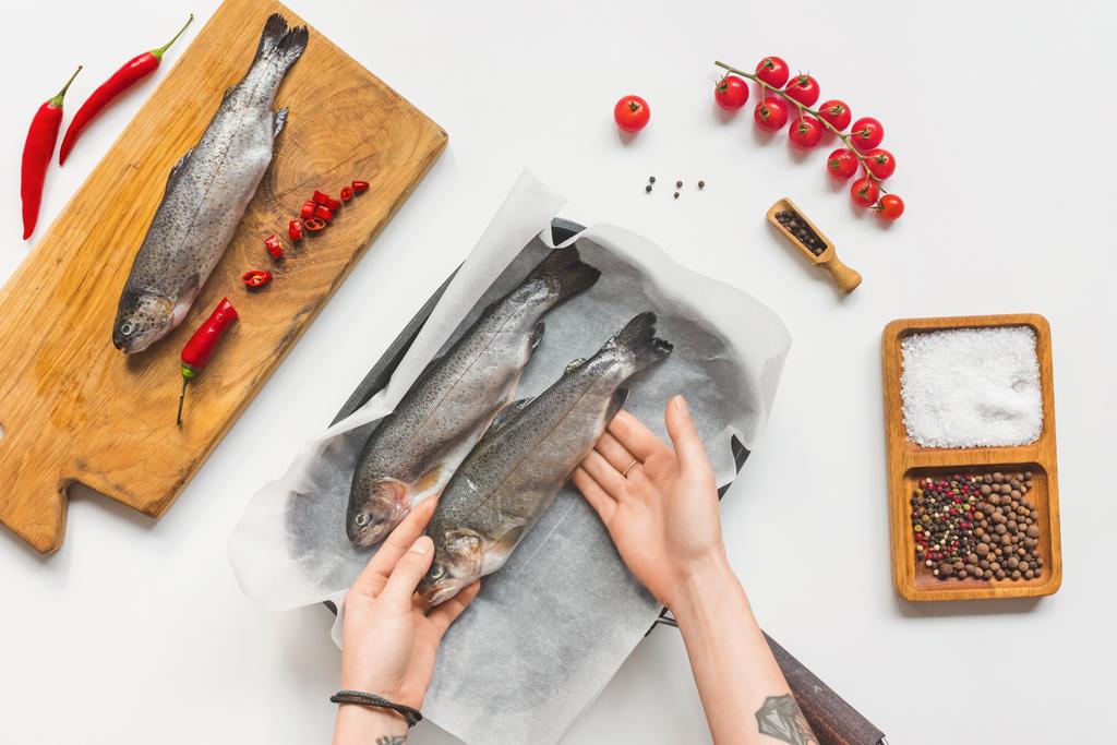 частковий вигляд жінки, що кладе неварену рибу в лоток з папером для випічки на білому столі з інгредієнтами
 - Фото, зображення