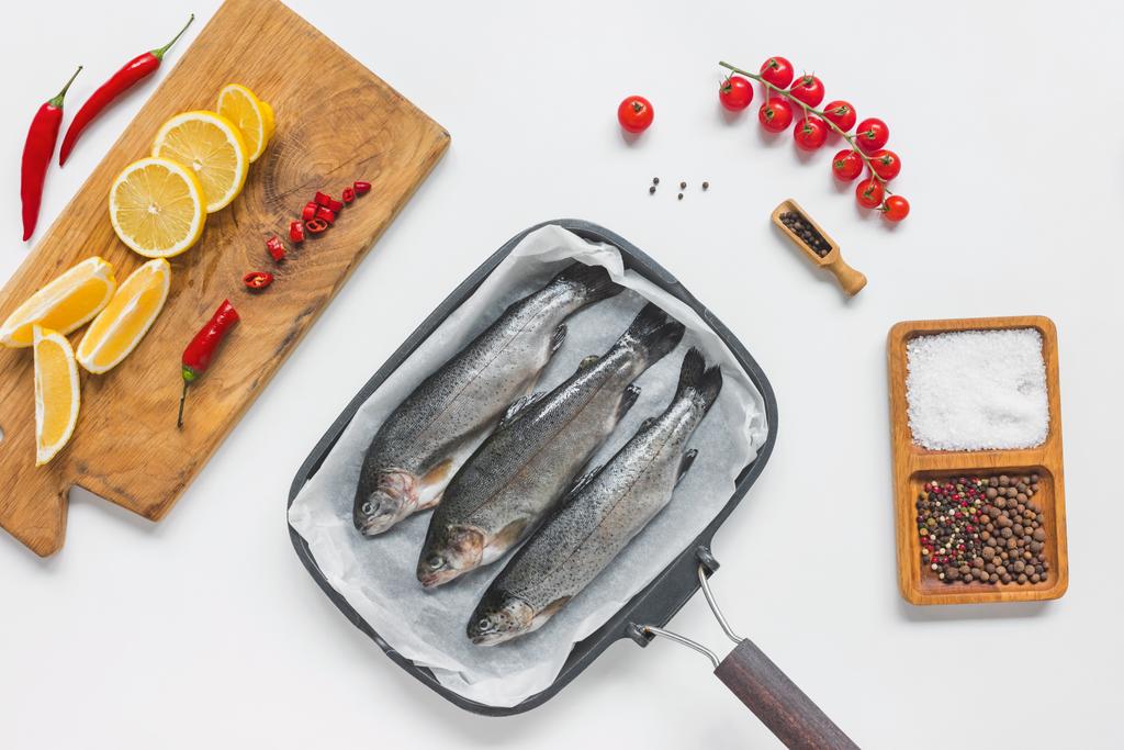вид на рыбу в подносе для выпечки в окружении различных ингредиентов на белом столе
 - Фото, изображение
