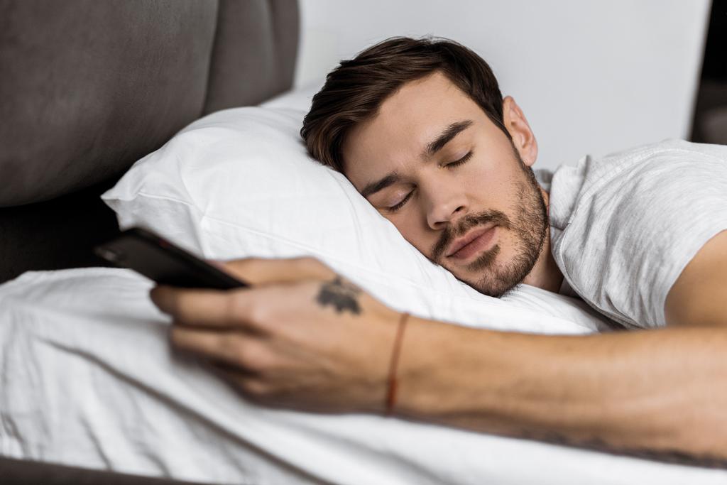 knappe bebaarde jongeman slapen in bed met de smartphone in de hand - Foto, afbeelding