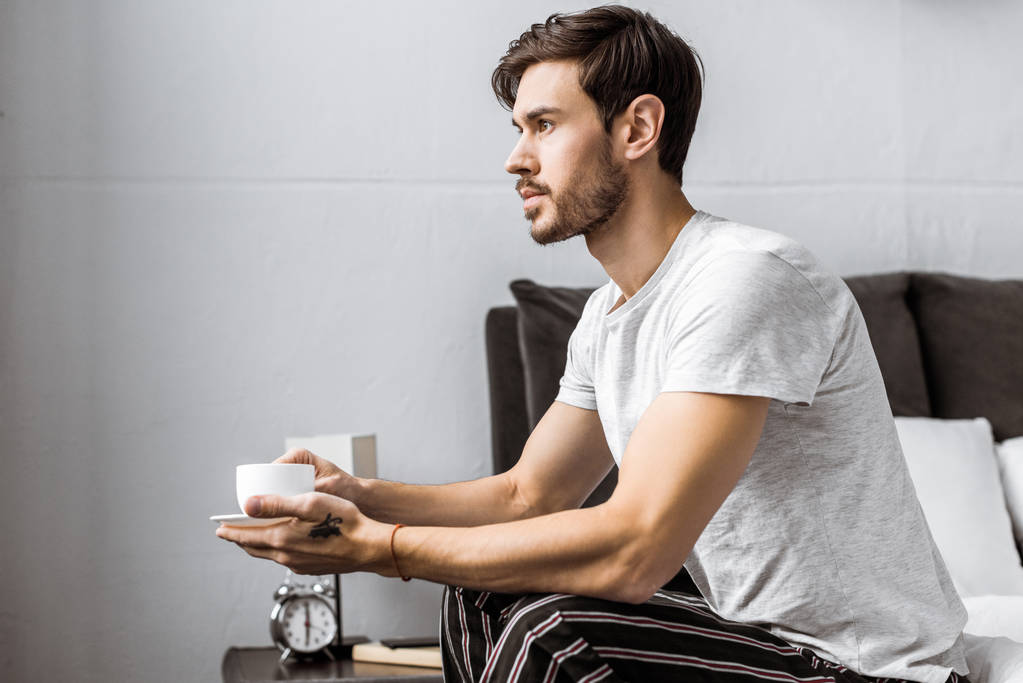 Seitenansicht eines jungen Mannes im Schlafanzug, der auf dem Bett sitzt und eine Tasse Kaffee hält - Foto, Bild