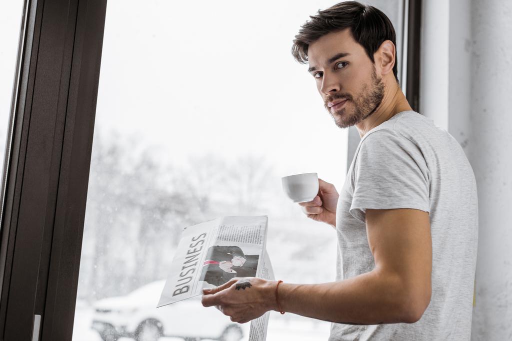 schöner junger Mann mit Tasse Kaffee und Zeitung, der in die Kamera schaut, während er am Fenster steht - Foto, Bild