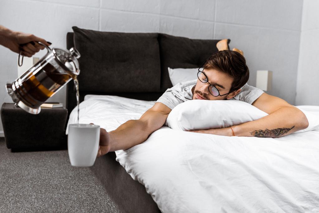 誰かがベッドの上に横たわると、カップを保持している眼鏡で眠い男中急須からお茶を注ぐのクロップ撮影 - 写真・画像