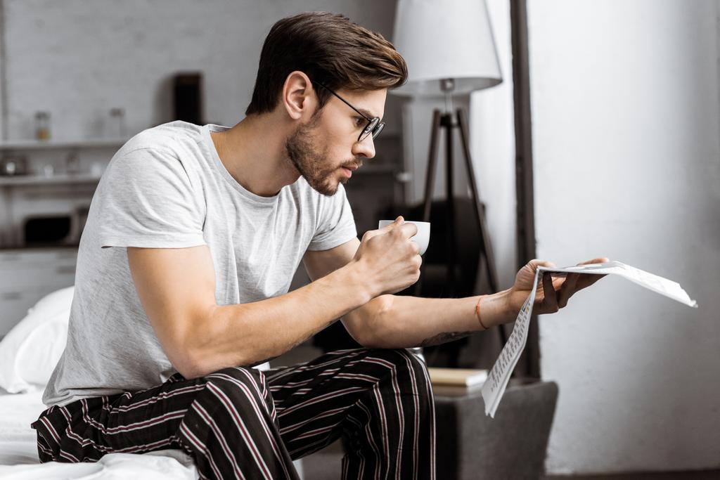 joven guapo en pijama y anteojos sosteniendo taza de café y leyendo el periódico en la mañana
 - Foto, imagen