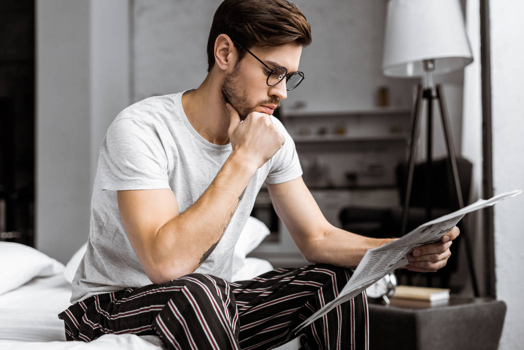 komea nuori mies pyjamassa ja silmälaseissa istuu sängyllä ja lukee sanomalehteä aamulla
 - Valokuva, kuva