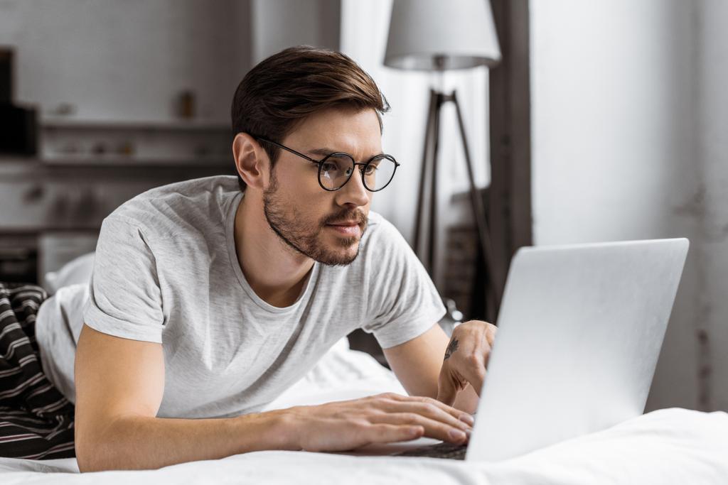 όμορφος νεαρός άνδρας με γυαλιά και πιτζάμες χρησιμοποιώντας το laptop στο κρεβάτι το πρωί - Φωτογραφία, εικόνα