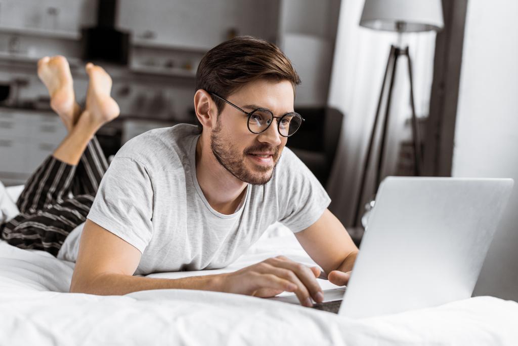 jeune homme souriant en lunettes et pyjama à l'aide d'un ordinateur portable sur le lit
 - Photo, image