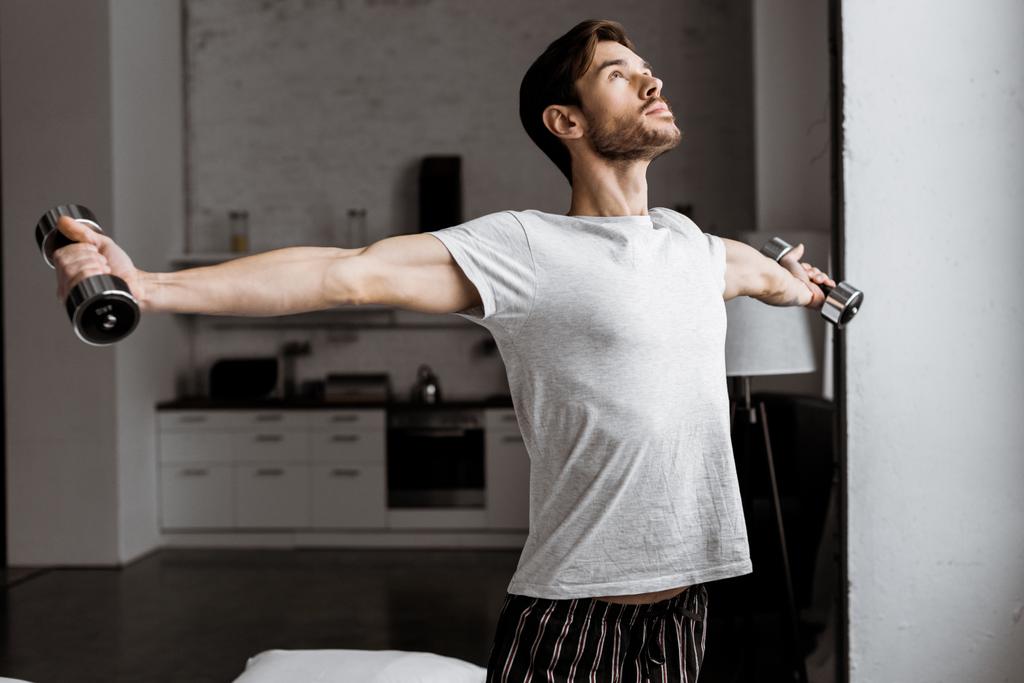 Красивый молодой человек в пижаме тренируется с гантелями и смотрит вверх на дом
  - Фото, изображение