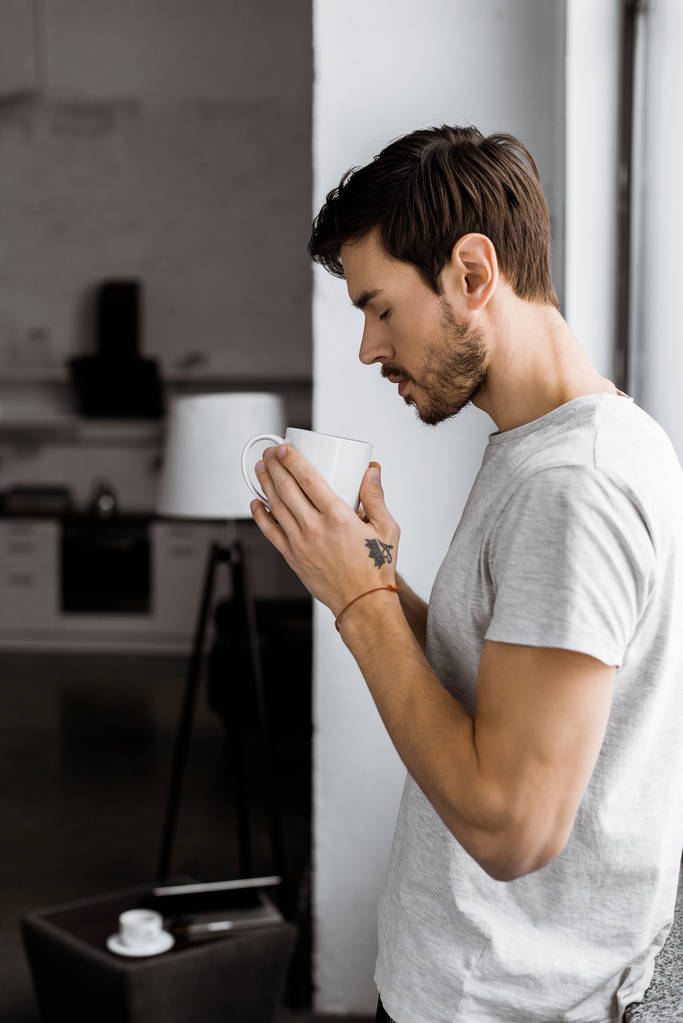 boční pohled pohledný mladý muž s hrnkem kávy opírající se o okno doma - Fotografie, Obrázek