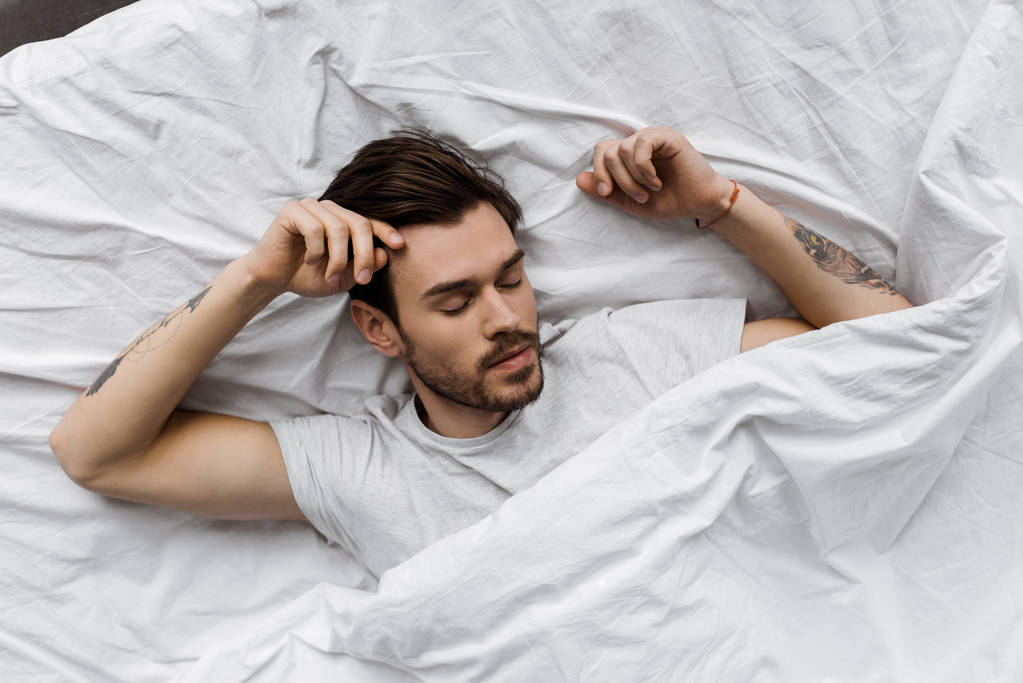 верхний вид красивого молодого человека, лежащего под белым одеялом в постели дома
 - Фото, изображение