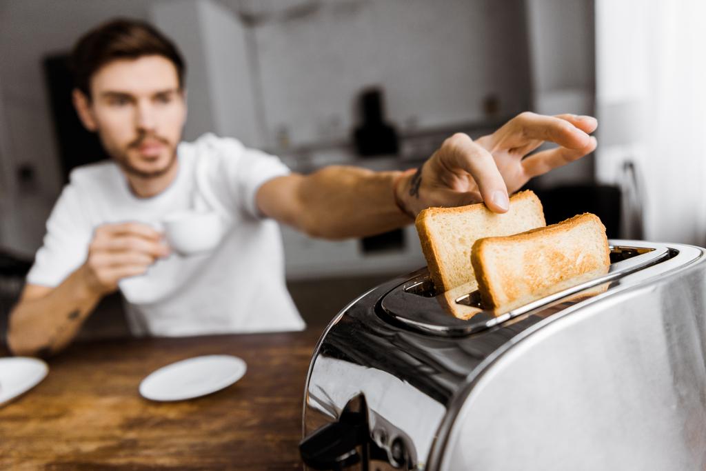 zbliżenie młody człowiek, picia kawy i biorąc od toster toast - Zdjęcie, obraz