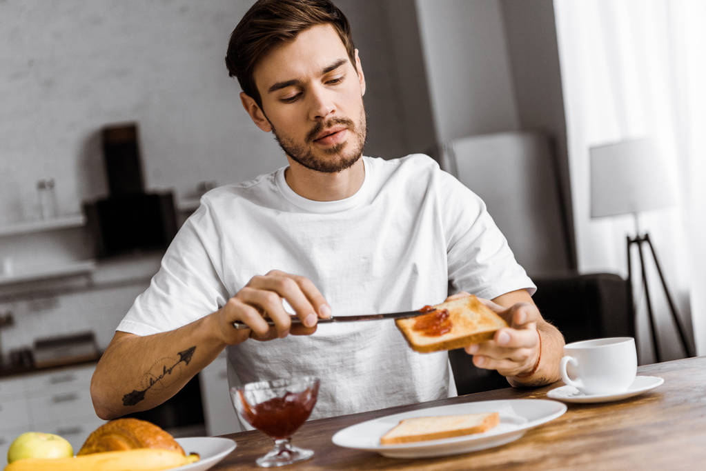 Красивый молодой человек, накладывающий варенье на тост в выходные утром дома
 - Фото, изображение