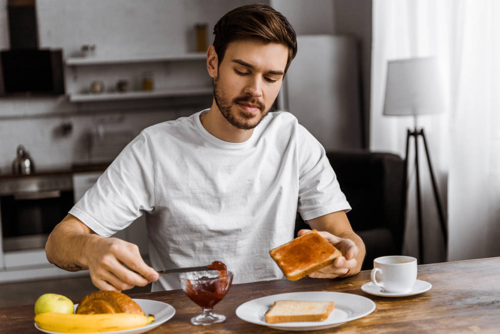 Привлекательный молодой человек наносит варенье на тосты дома
 - Фото, изображение