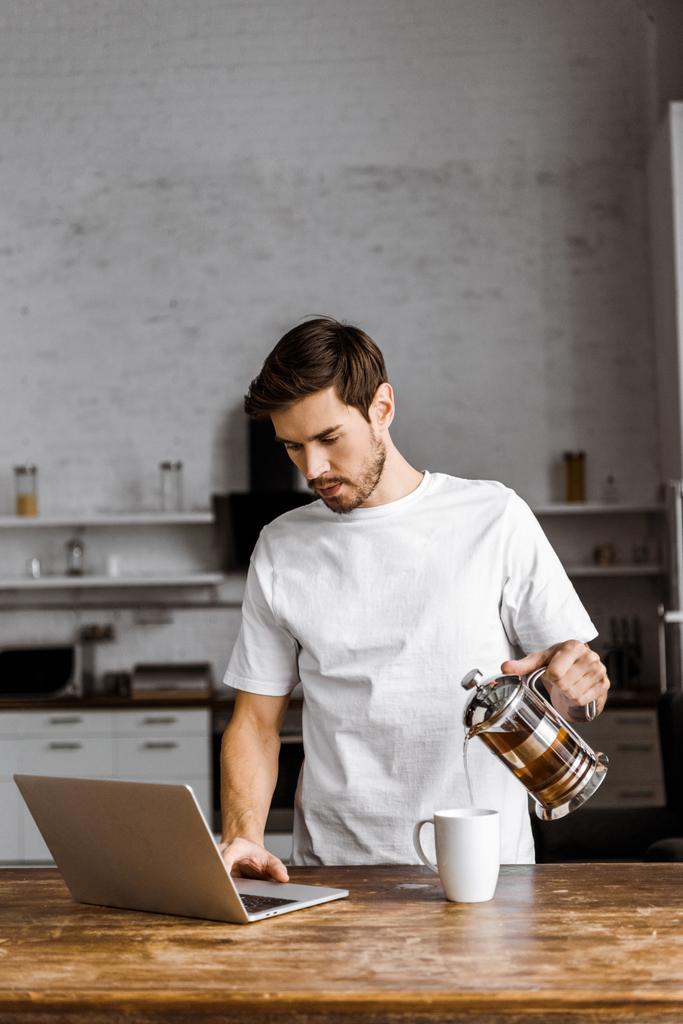 apuesto joven freelancer con taza de té, croissant y portátil trabajando en la cocina en casa
 - Foto, Imagen