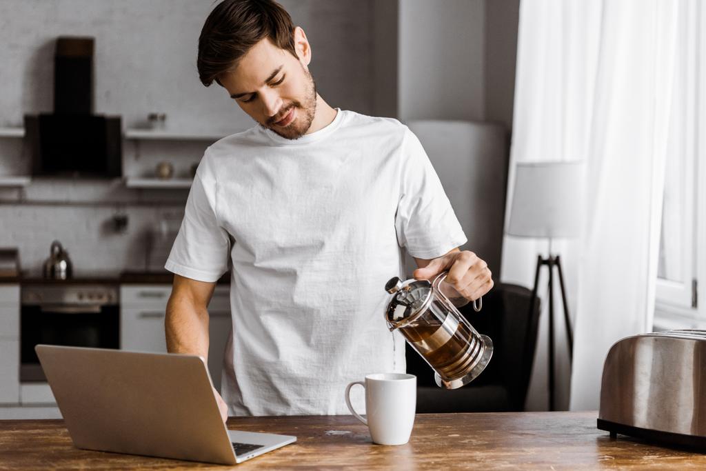 atraktivní mladá freelancer s notebookem nalil čaj do šálku na kuchyni doma - Fotografie, Obrázek