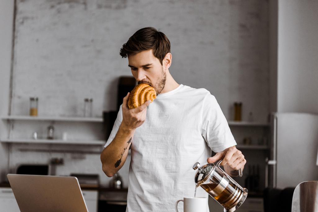 handsome young freelancer with cup of tea, croissant and laptop working on kitchen at home - Photo, Image