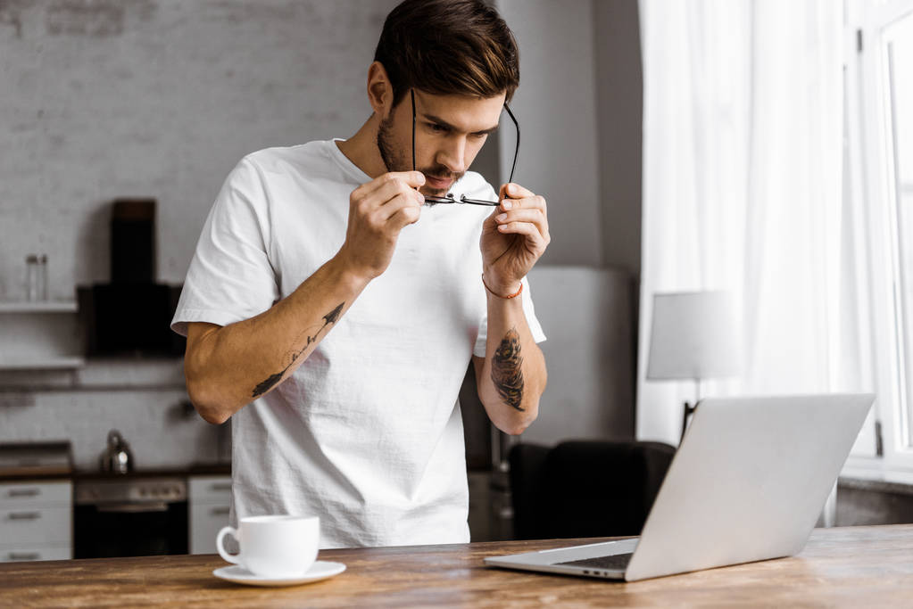 όμορφος νέος ελεύθερος επαγγελματίας σε γυαλιά με καφέ και φορητό υπολογιστή στην κουζίνα στο σπίτι - Φωτογραφία, εικόνα