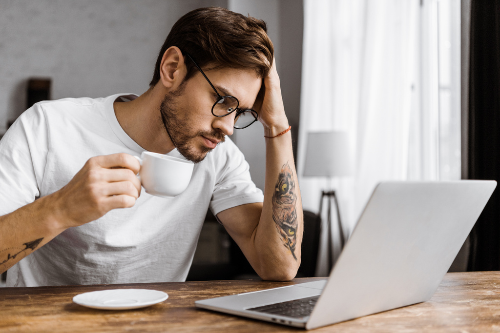 ylityöllistetty nuori freelancer kahvia katsomalla kannettavan tietokoneen näytön kotona
 - Valokuva, kuva