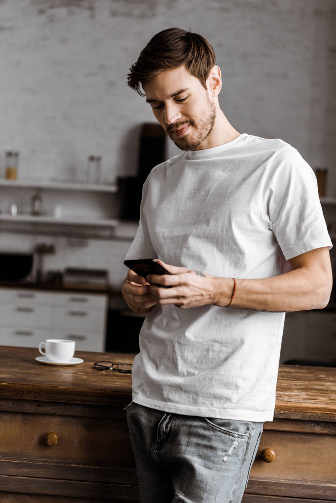 joven guapo usando teléfono inteligente en la cocina en casa
 - Foto, Imagen