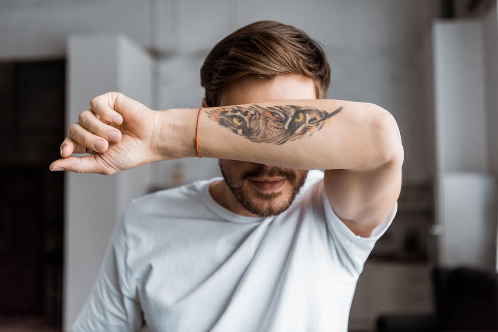 handsome young man covering face with arm with tattooed tiger eyes at home - Photo, Image