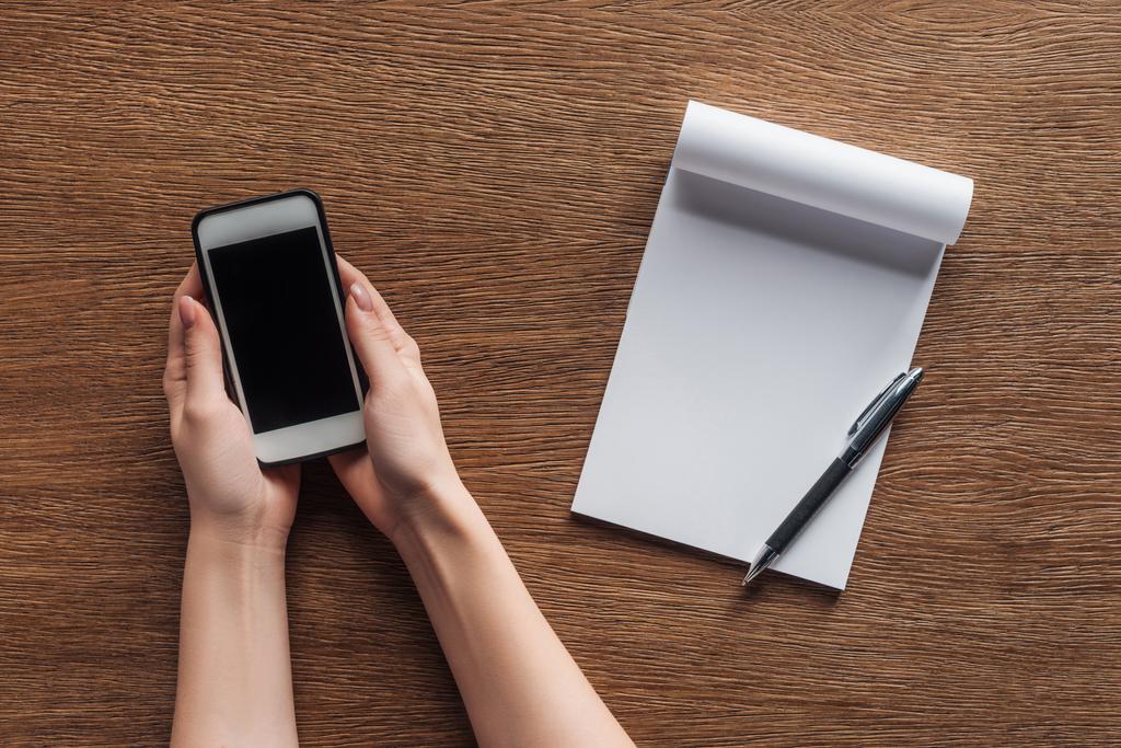 Teilansicht der Person, die Smartphone mit leerem Bildschirm, Stift, Notizbuch mit Kopierraum auf Holzgrund hält - Foto, Bild