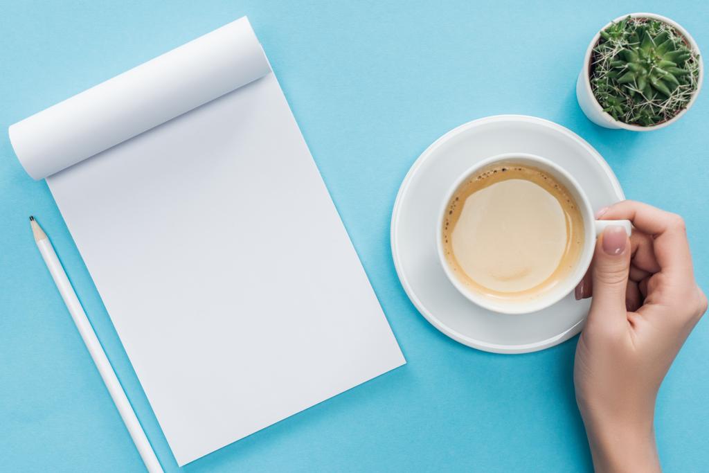 fincan kahve, boş defter ve kalem mavi arka plan üzerinde tutan kişinin kırpılmış görünümü - Fotoğraf, Görsel