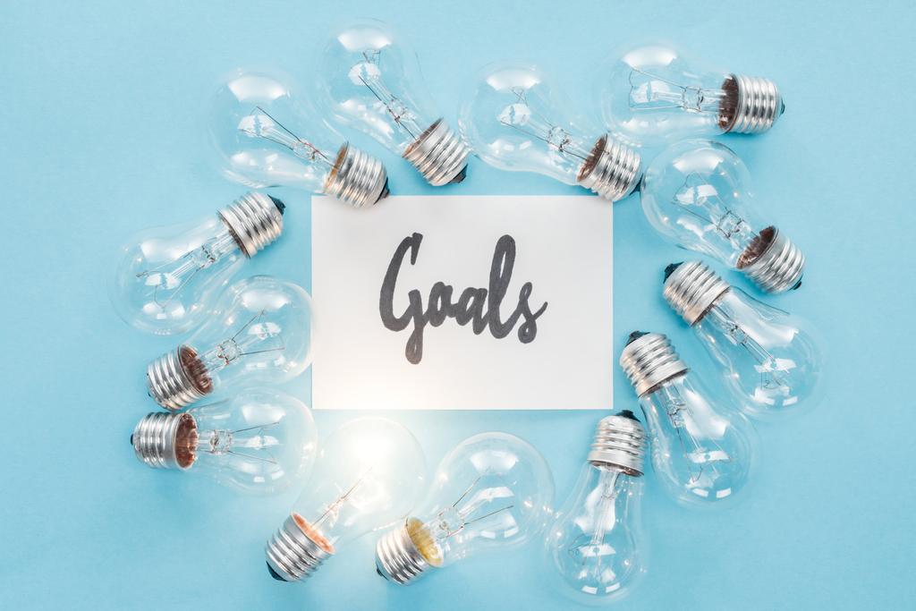 top view of 'goals' word written in cursive on card surrounded by circle of light bulbs on blue background, goal setting concept - Photo, Image