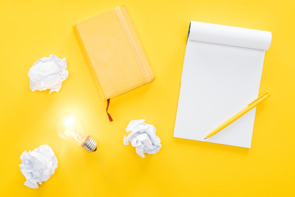top view of blank notebook, crumbled paper balls and glowing light bulb on yellow background, having new ideas concept - Photo, Image