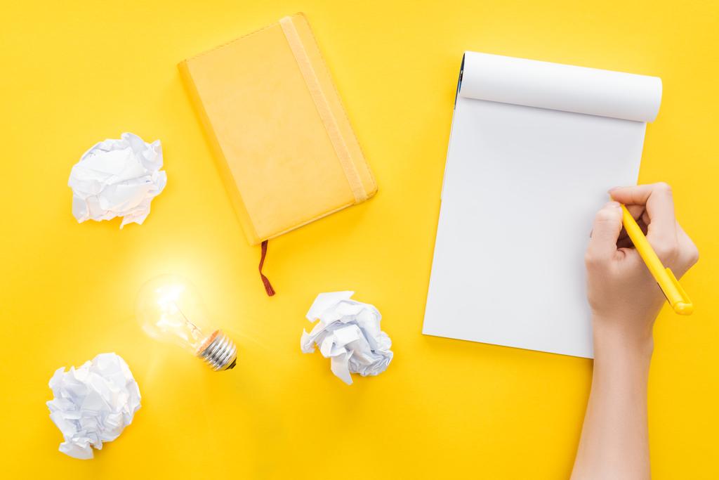 abgeschnittene Ansicht einer Frau, die in leeres Notizbuch schreibt, zerbröselte Papierkugeln und glühende Glühbirne auf gelbem Hintergrund, mit neuen Ideen Konzept - Foto, Bild