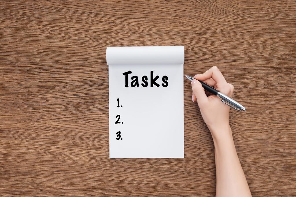 Ausgeschnittene Ansicht einer Frau, die Wort "Aufgaben" in Notizbuch auf Holzgrund schreibt - Foto, Bild