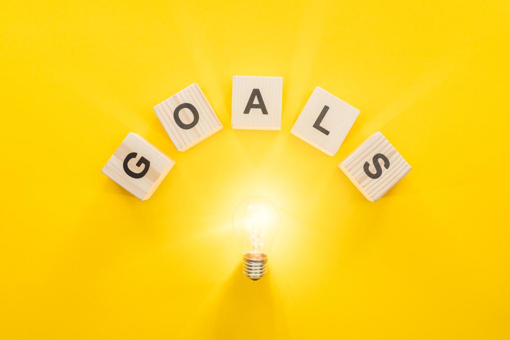 top view of glowing light bulb under 'goals' word made of wooden blocks on yellow background, goal setting concept - Photo, Image