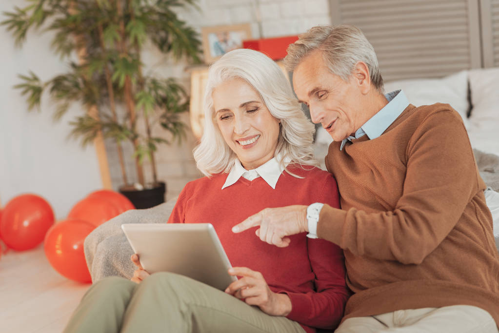 Интересный контент. Счастливая пожилая супружеская пара сидит в гостиной и смотрит фотографии на планшете, радостно улыбаясь и указывая на экран планшета
 - Фото, изображение
