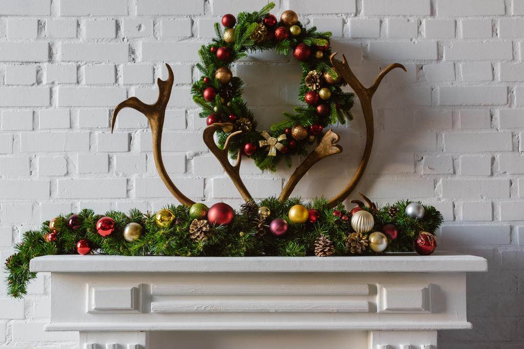 corona de Navidad con decoraciones y cuernos de ciervo sobre chimenea mantel
 - Foto, Imagen