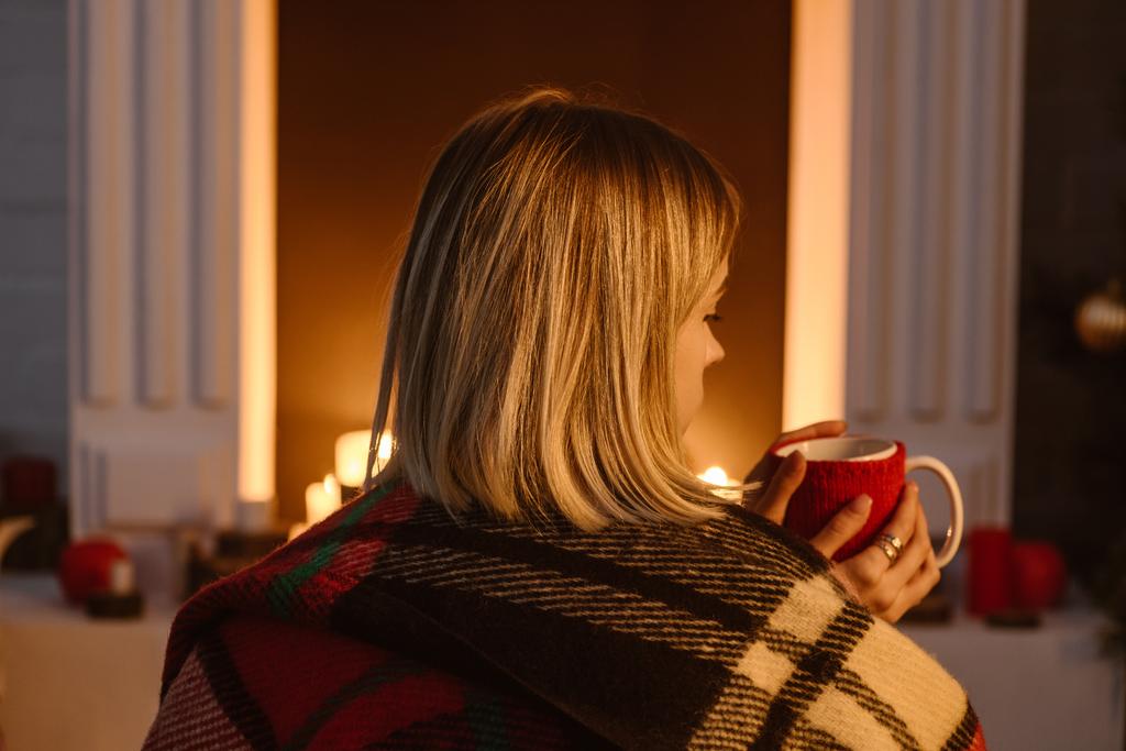 Achteraanzicht van de vrouw in geruite deken zitten in de buurt van open haard en houden van kopje koffie thuis - Foto, afbeelding