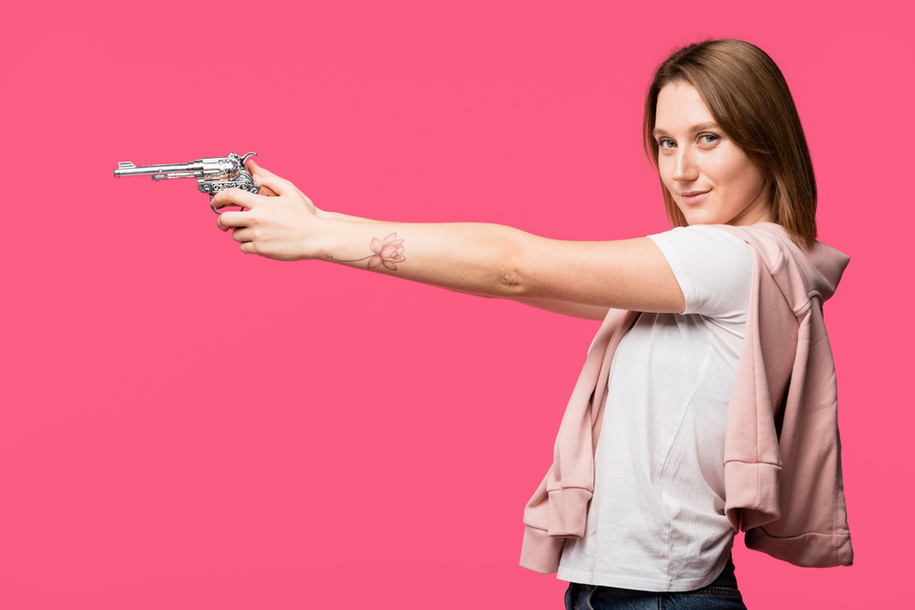 Widok z boku młodej kobiety, trzymając rewolwer i uśmiecha się do kamery na różowym tle - Zdjęcie, obraz