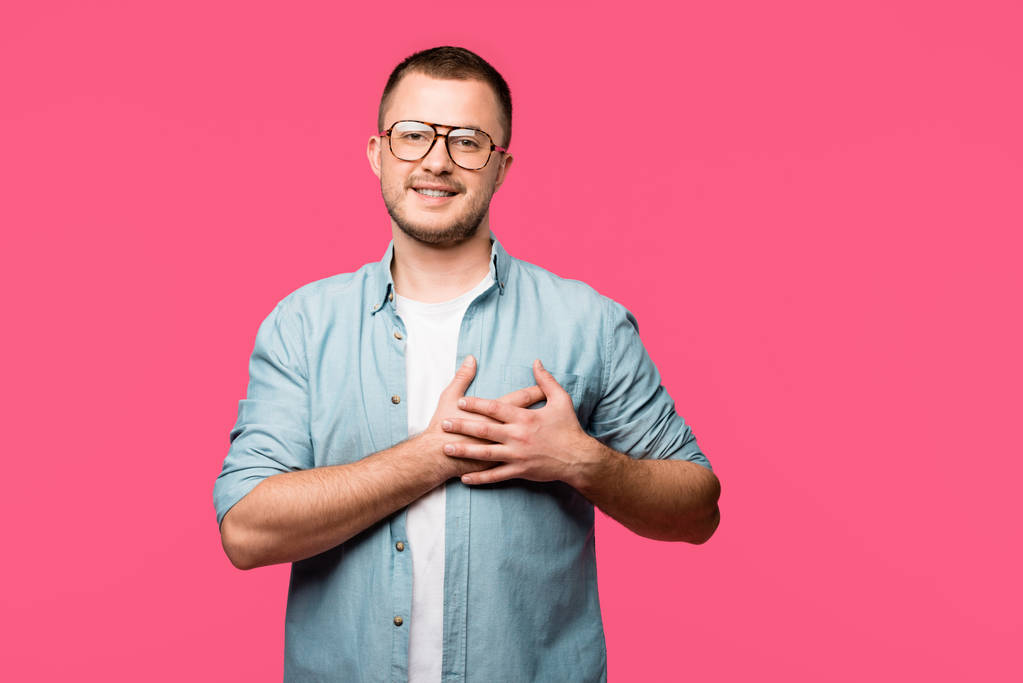 onnellinen nuori mies silmälaseissa pitäen kädestä sydämen yläpuolella ja hymyillen kameralle eristettynä vaaleanpunaiseen
 - Valokuva, kuva