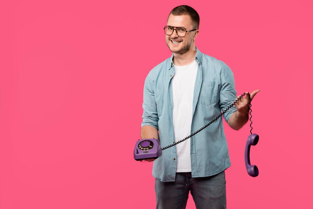 Красивый молодой человек в очках, держащий фиолетовый ротационный телефон и улыбающийся в камеру, изолированную на розовый
 - Фото, изображение