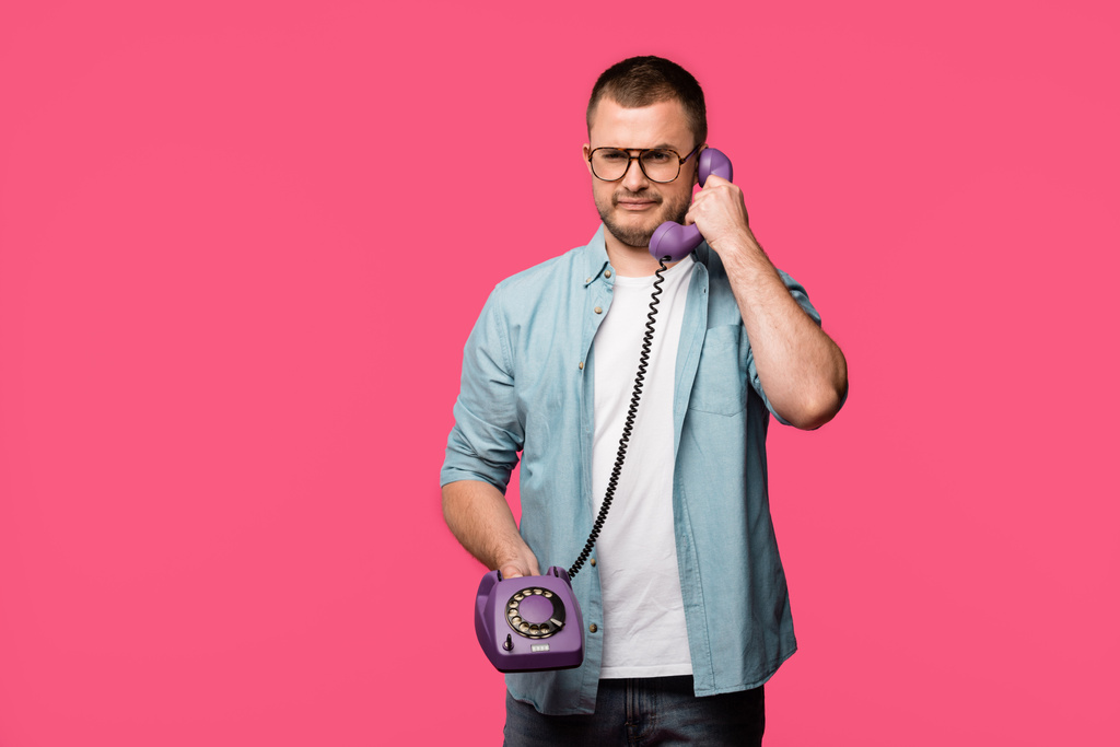 frowning young man talking by vintage telephone and looking at camera isolated on pink - Photo, Image