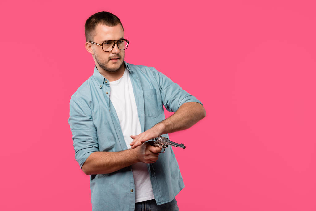 young man in eyeglasses reloading revolver and looking away isolated on pink - Photo, Image