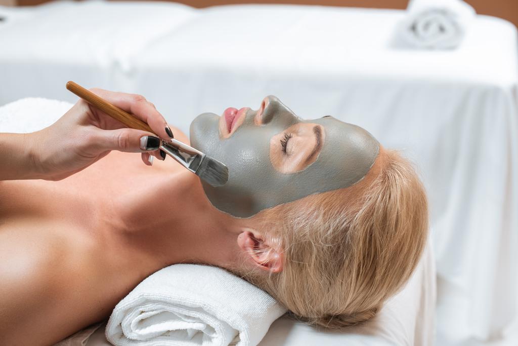 κοντινό πλάνο αισθητικός εφαρμόζει μάσκα αργίλου με πινέλο στο πρόσωπο της γυναίκας - Φωτογραφία, εικόνα