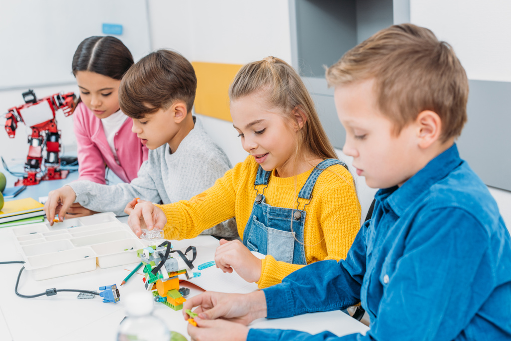 концентрированные дети, сидящие за столом и делающие роботов в классе по обучению основам
 - Фото, изображение