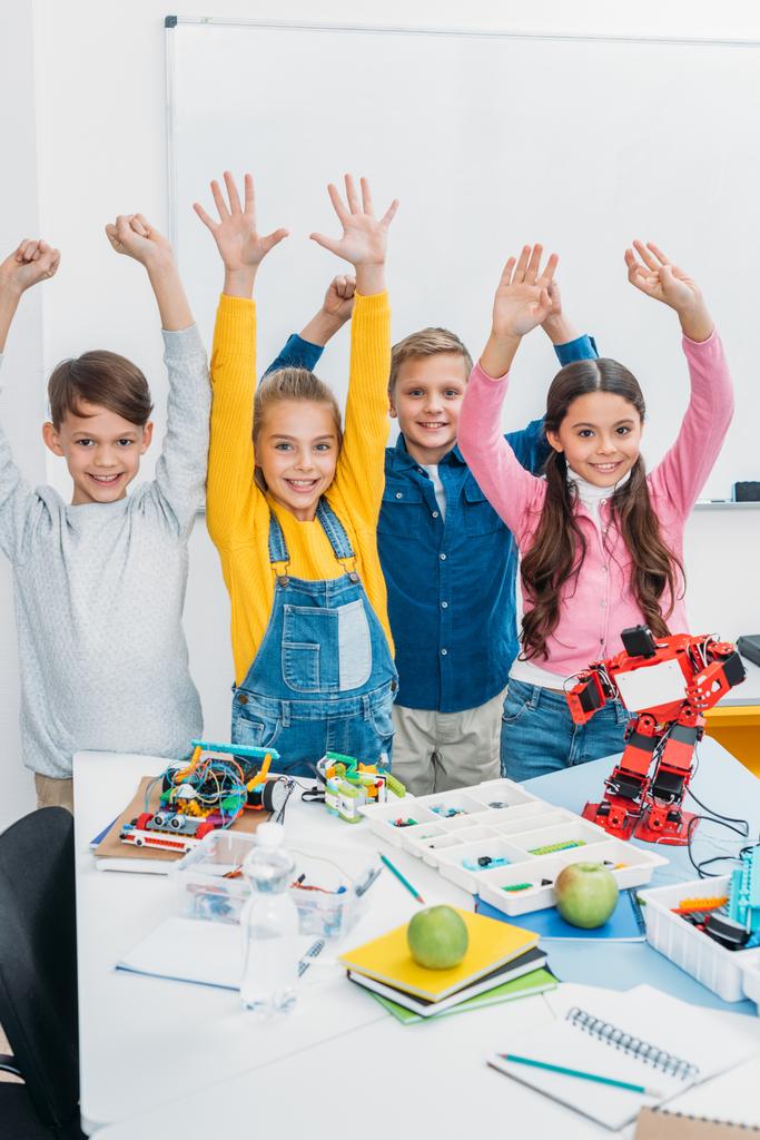 glückliche Schulkinder mit erhobenen Händen am Schreibtisch mit Plastikrobotern und Details in der Stammklasse - Foto, Bild