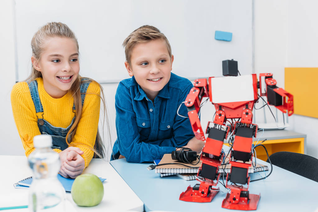 Lächelnde Schulkinder sitzen am Schreibtisch und betrachten einen roten Plastikroboter in der Klasse - Foto, Bild