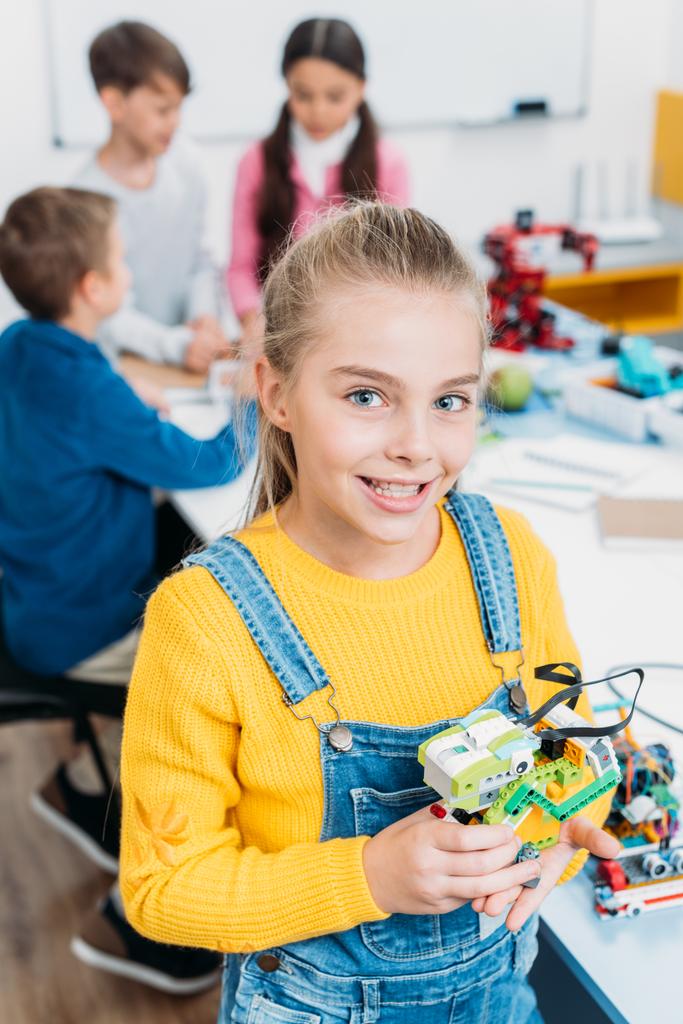 preteen schoolgirl holding multicolored robot and looking at camera in classroom - Photo, Image
