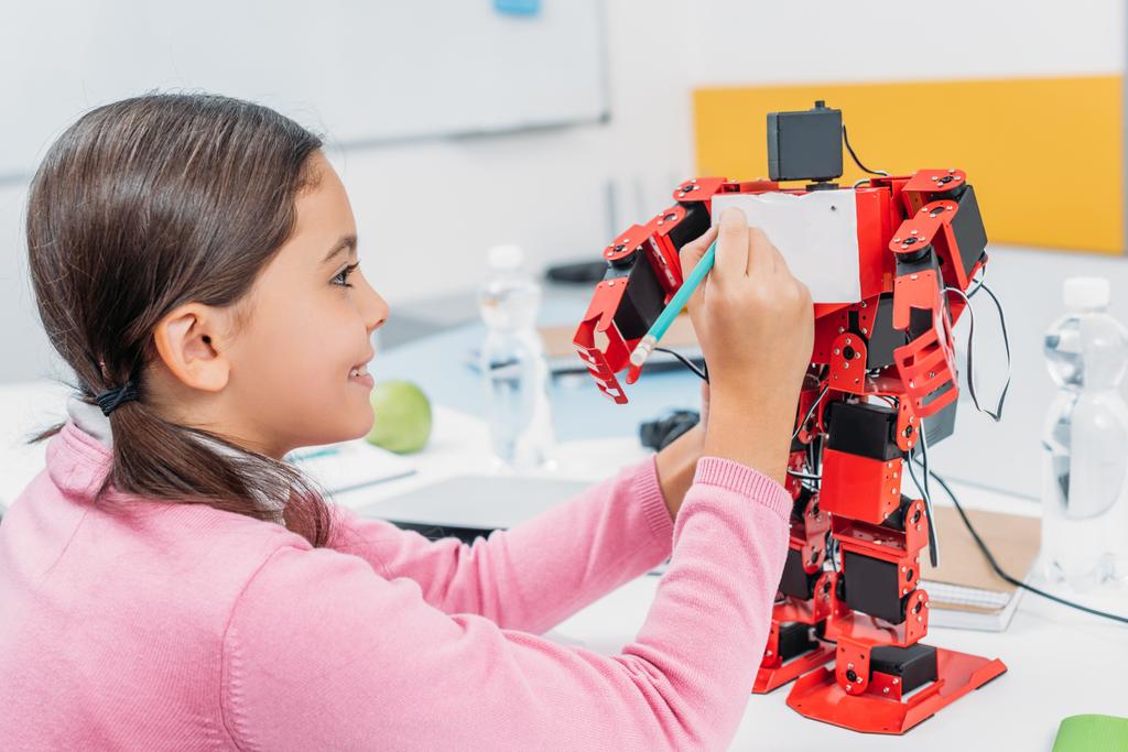 s úsměvem školačka psaní tužkou na hrudi červené robota v kmenové třídě - Fotografie, Obrázek