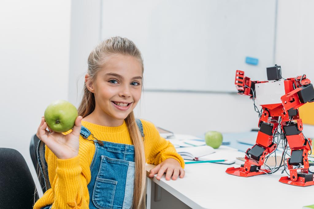 Lächelndes Schulmädchen sitzt mit Robotermodell am Tisch im Klassenzimmer, hält Apfel in der Hand und blickt in die Kamera - Foto, Bild